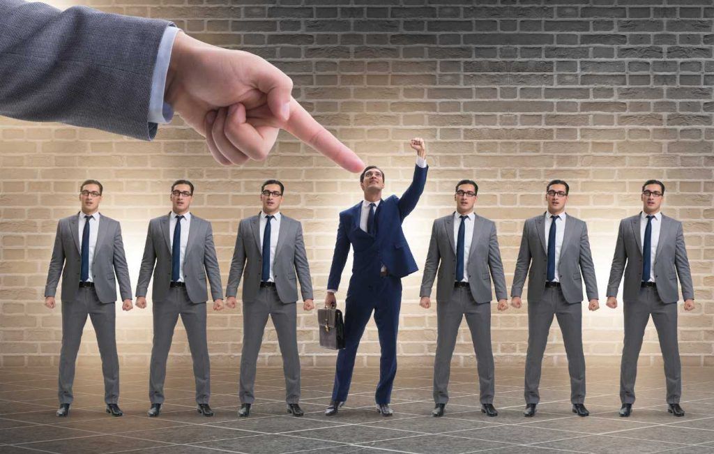 Large hand points at a businessman raising his arm, standing out from other men in suits.