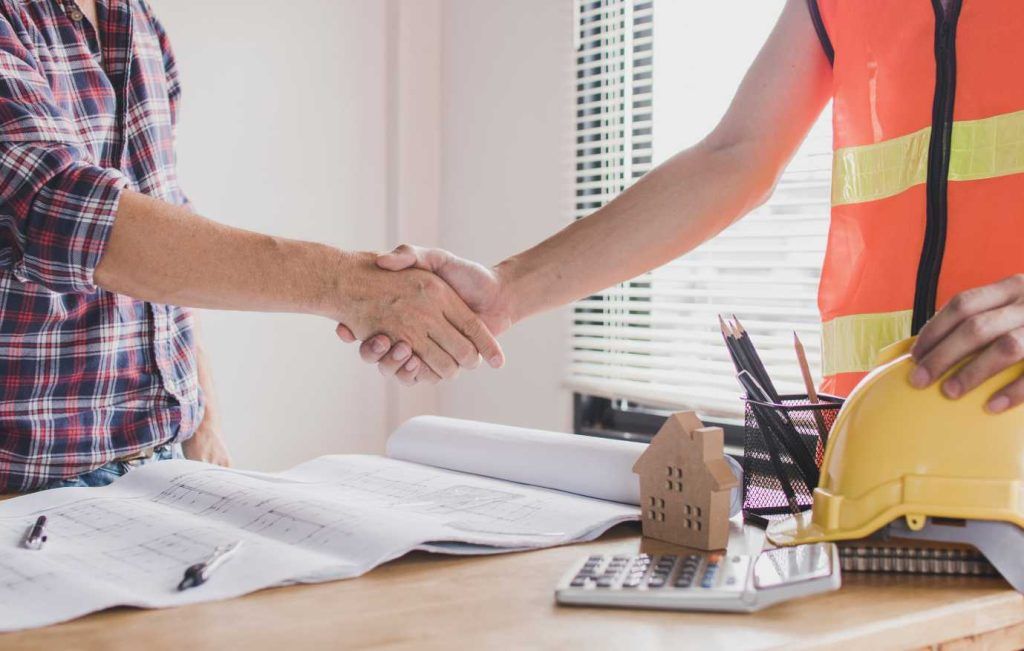 Two people shaking hands over a desk with blueprints, a model house, and a calculator. One wears a hard hat.