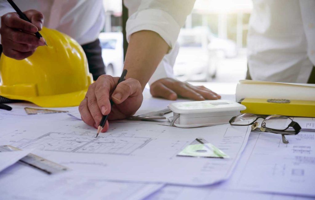 Hands of architects working on blueprints with yellow hardhat nearby.