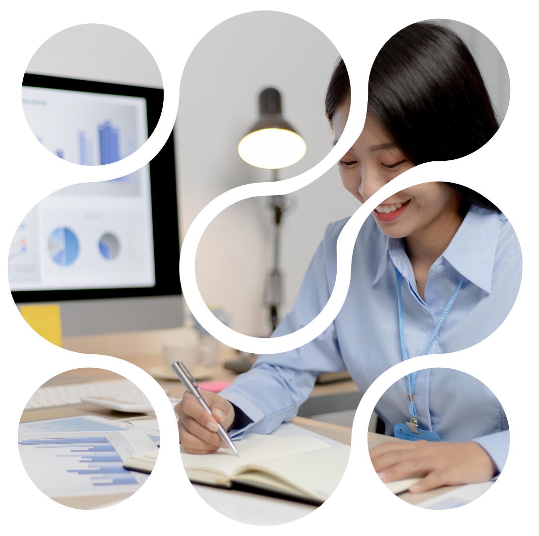 Woman in blue shirt writing in a notebook, working at her desk with a computer and documents.