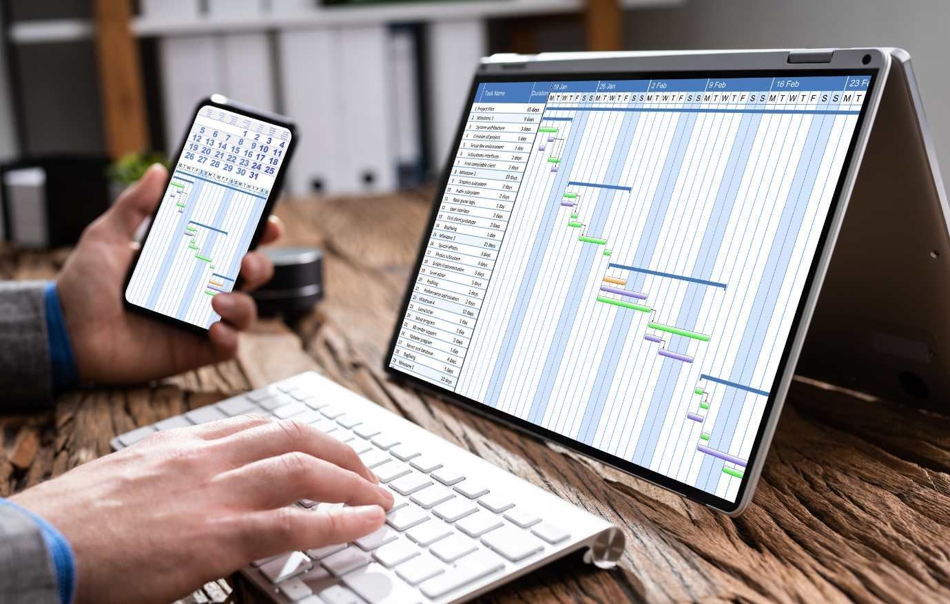 Person uses phone and laptop, both displaying a project schedule, while typing on a keyboard at a desk.