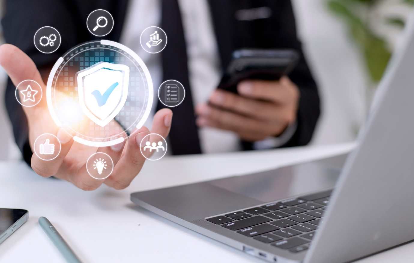 Person holding a digital shield icon with checkmark, near laptop and phone, symbolizing cybersecurity.