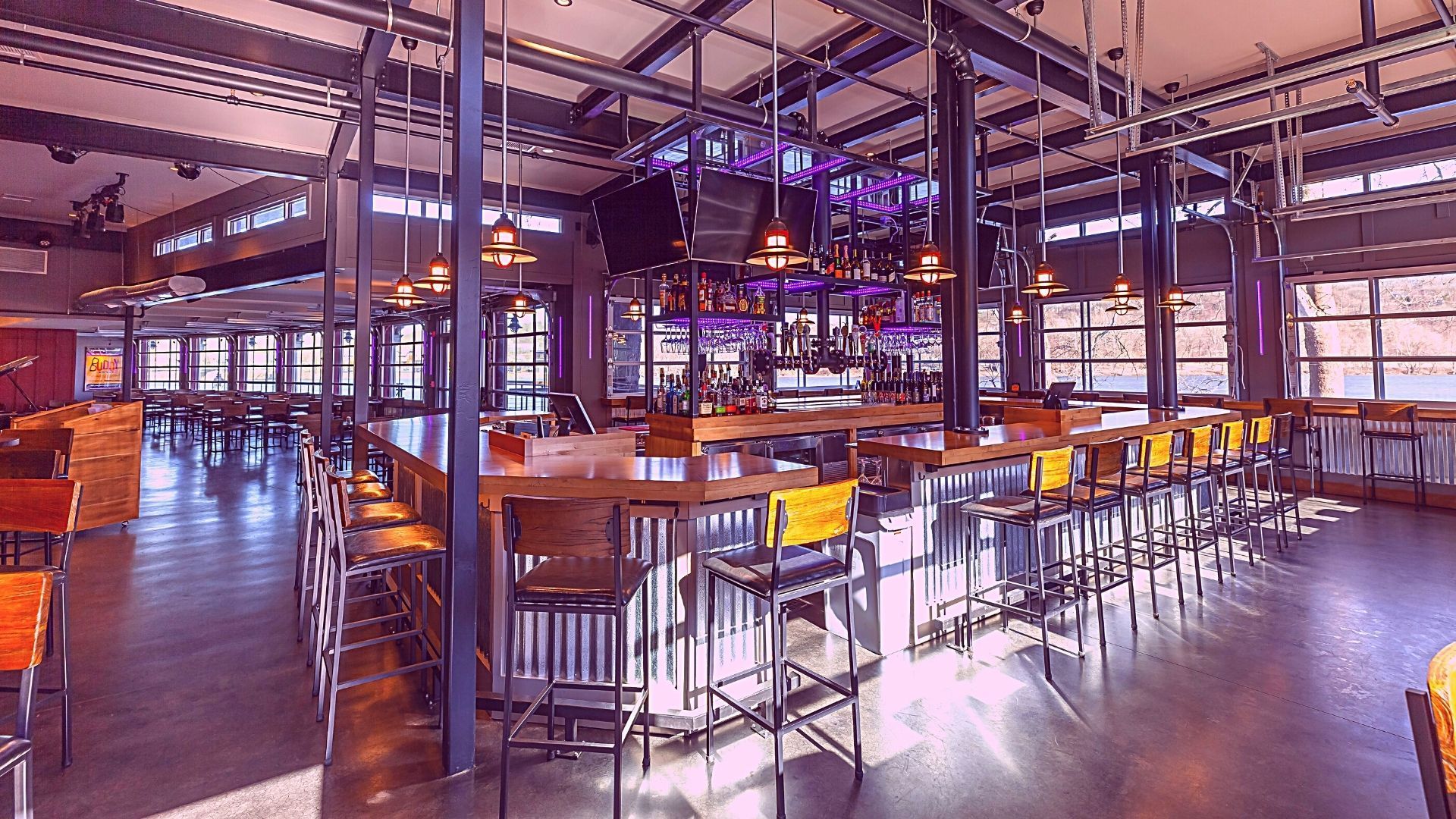 A modern bar with a long wooden counter, metal stools, and large windows.