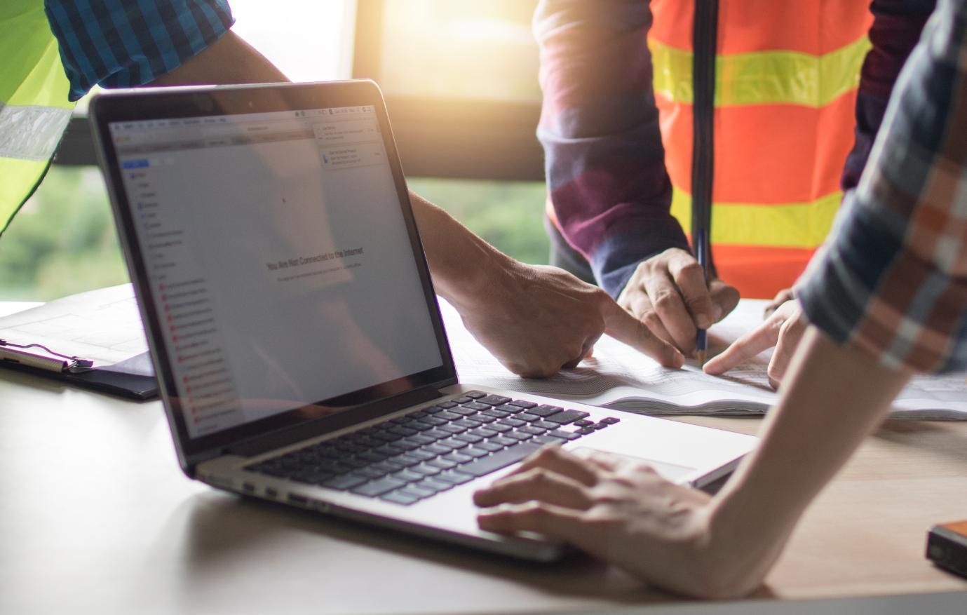 People in construction attire working together at a laptop and papers on a table.