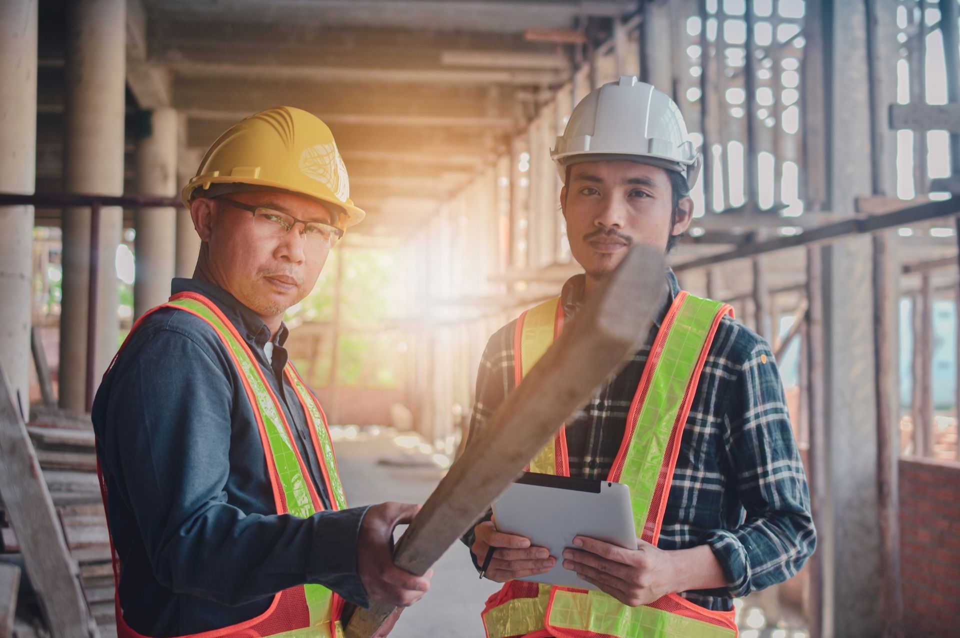 Two construction workers in hard hats and safety vests on a construction site, one holding a wooden beam.