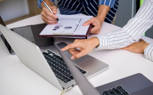 Two people reviewing documents and pointing at a laptop, collaborative office setting.
