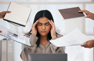 Woman with hands on her temples, overwhelmed by papers and devices in a business setting.