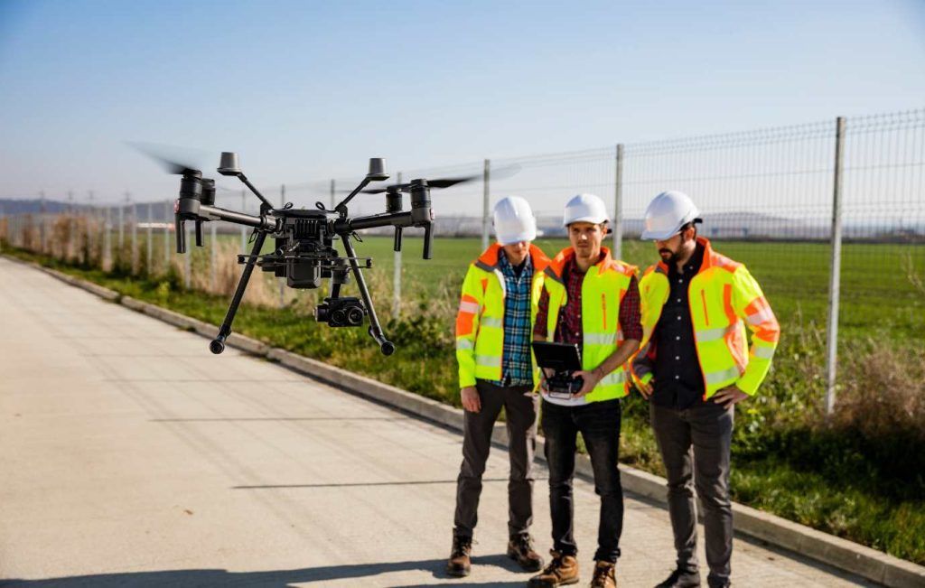 Three construction workers in safety vests and helmets operate a drone outdoors.