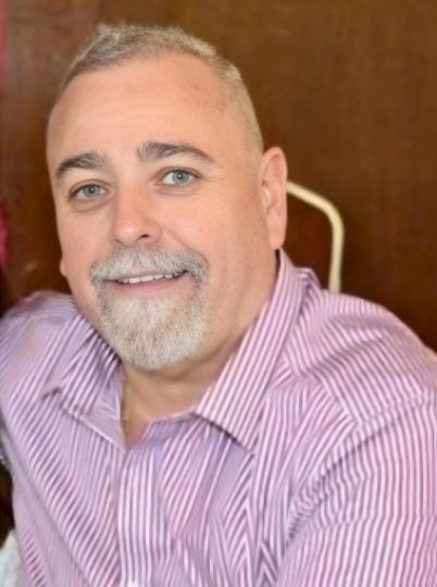 Man with short gray hair, goatee, and pink striped shirt, smiling.
