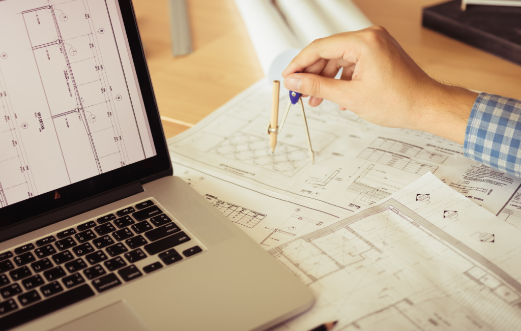 Person using a compass on architectural plans next to a laptop on a wooden desk.