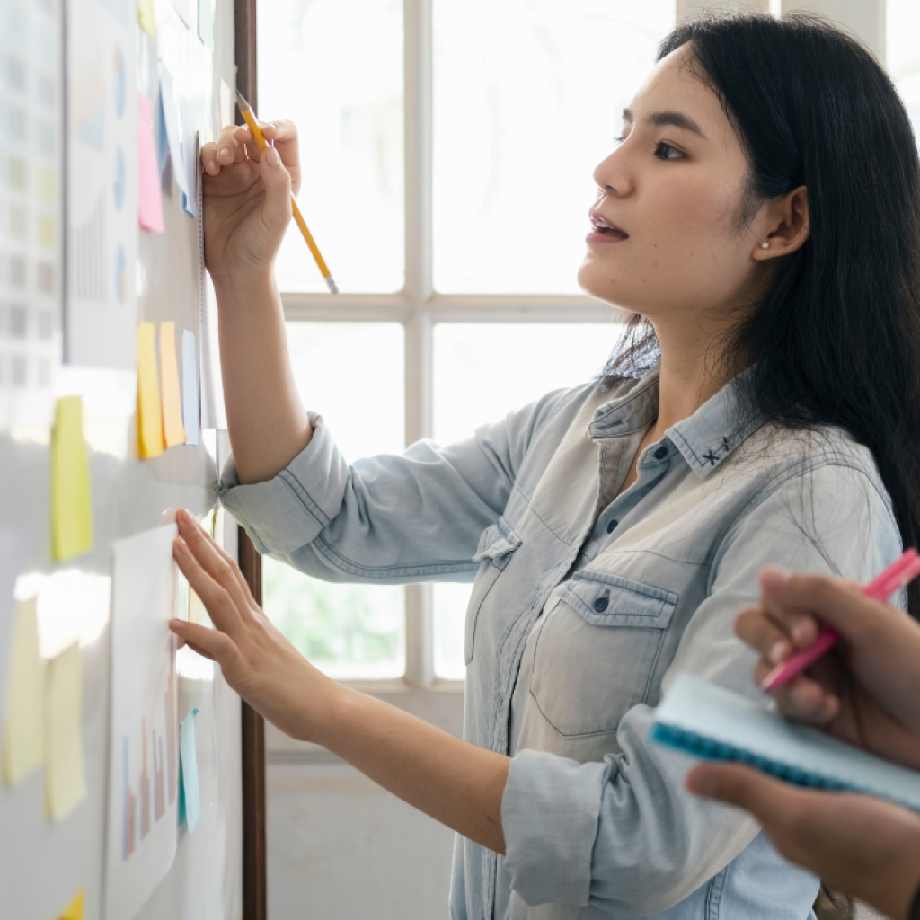 Woman writes on a whiteboard covered with sticky notes, working with a person holding a notepad.