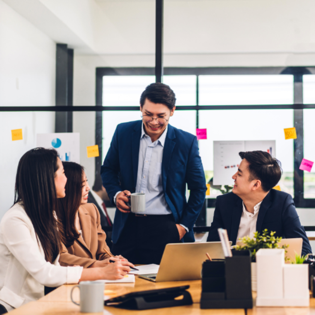 Business team in a meeting; man standing, smiling. Modern office with charts and sticky notes.