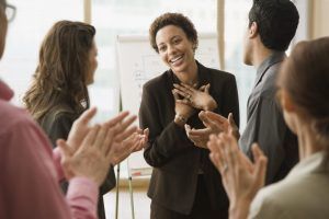 Smiling businesswoman receiving applause from colleagues in an office setting.