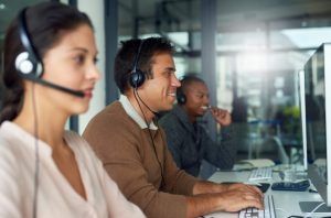 Three call center workers wearing headsets, smiling, and working on computers.