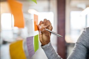 Person writing on orange sticky note on glass, brainstorming ideas.