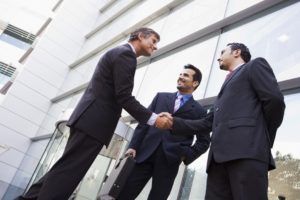Three men in suits shaking hands outside a building, smiling.