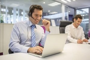 Man with headset smiles while typing on a laptop in an office; another man writes at a desk nearby.