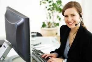 Woman wearing a headset smiles while typing at a computer in an office setting.
