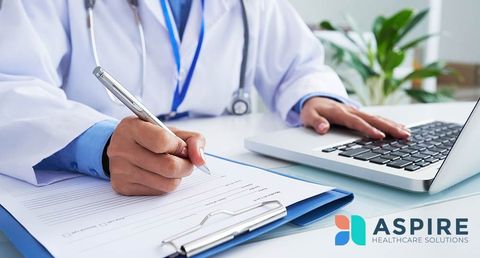A doctor is writing on a clipboard while using a laptop computer.