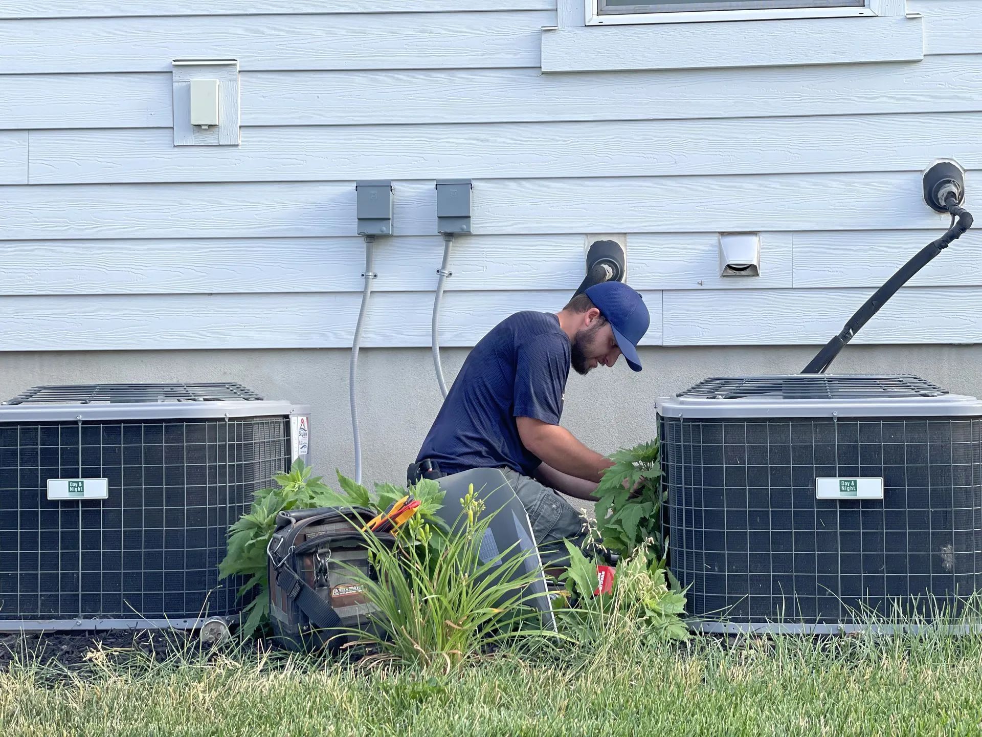 A person in a blue shirt and hat working on two air conditioning units near a light gray house.