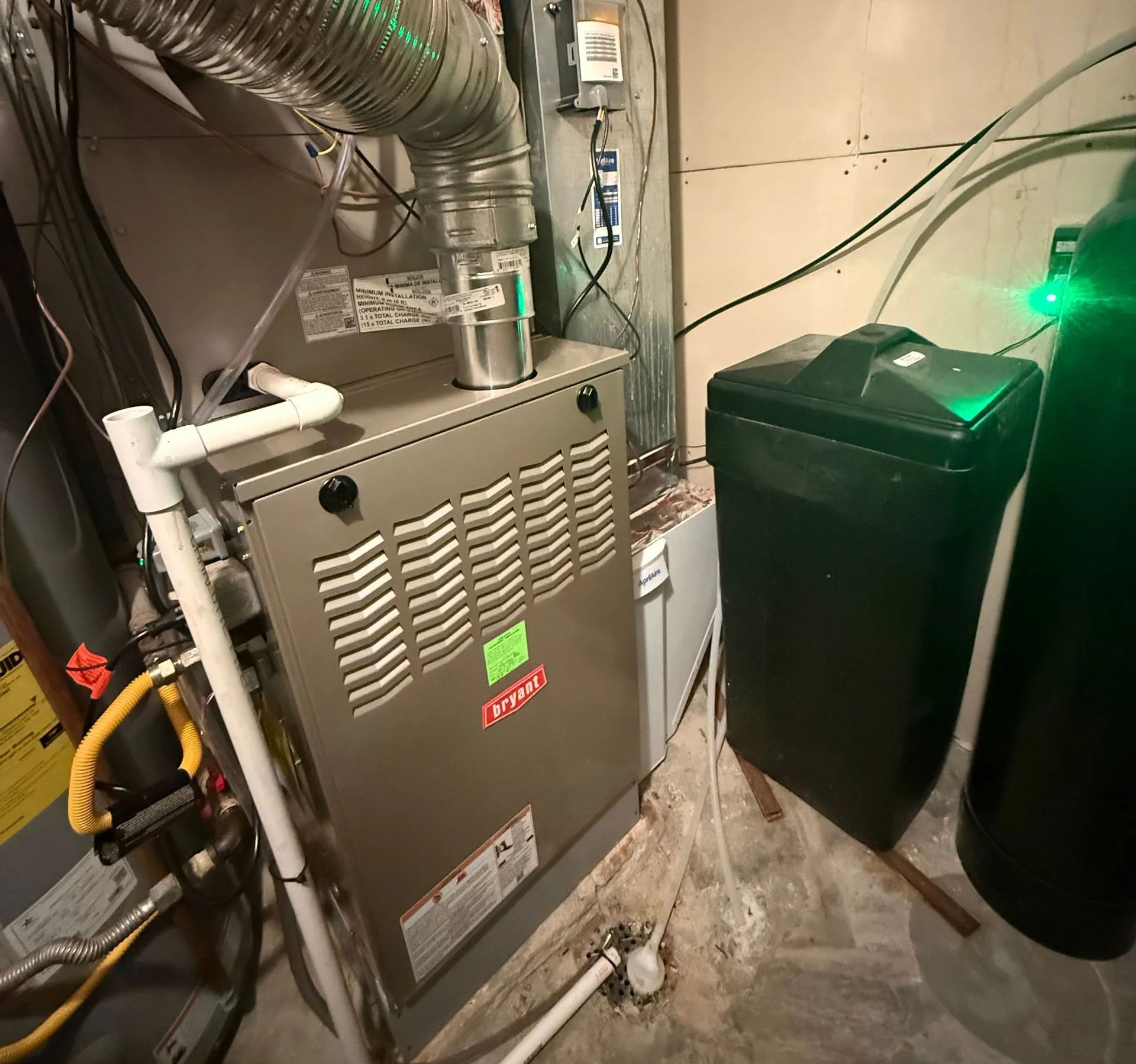 Basement HVAC furnace beside black trash bin and water heater, with ductwork and pipes visible
