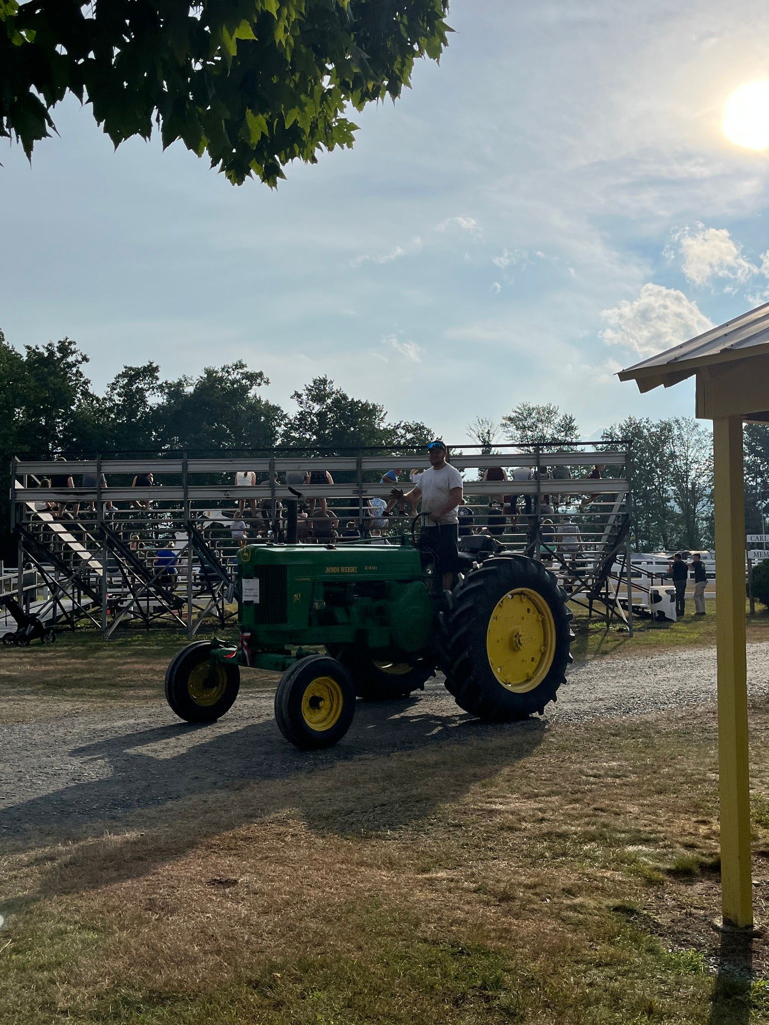 Person driving a green vintage tractor in a sunlit outdoor arena with bleachers in the background