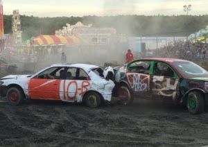 Two damaged race cars collide in a dusty demolition derby track with smoke and spectators behind them