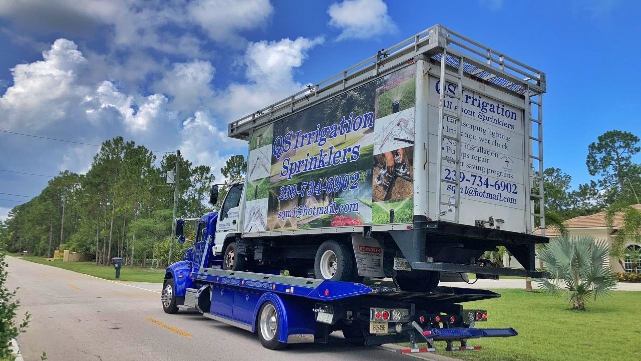 Blue flatbed tow truck hauling a large white trailer on a sunny roadside with trees and blue sky.