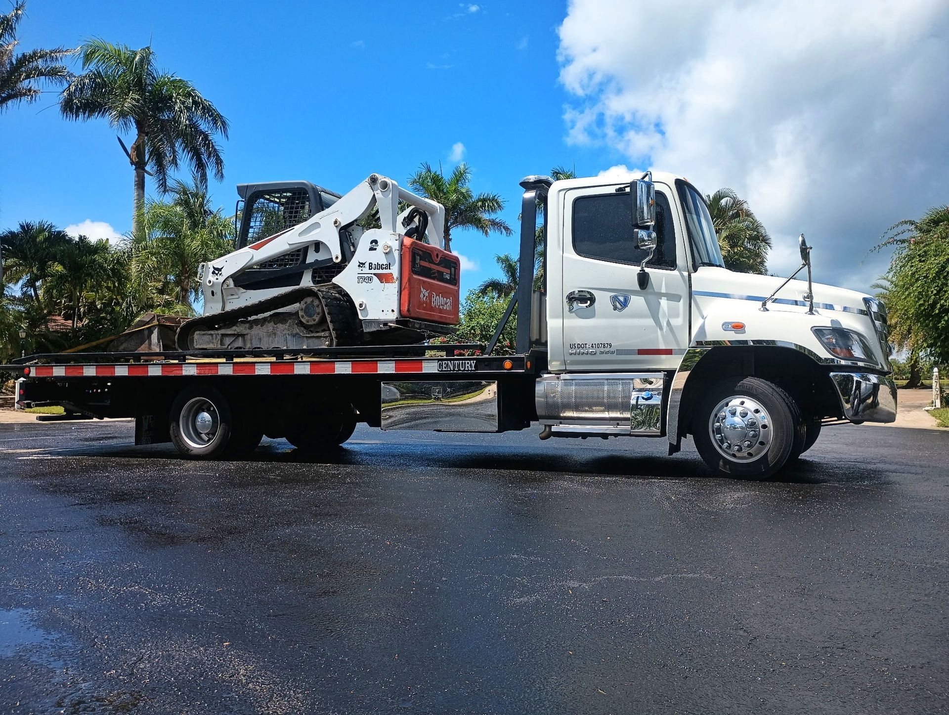 Tow truck carrying a white skid-steer loader on a flatbed in a wet parking lot under a blue sky
