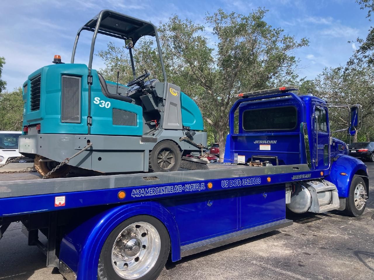 Blue flatbed tow truck carrying a turquoise road roller on a sunny roadside