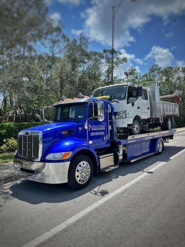Blue tow truck carrying a white pickup on a flatbed trailer beside a road lined with trees