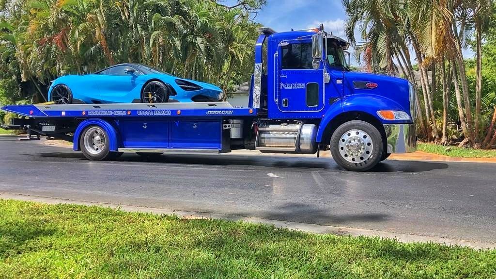 Blue tow truck hauling a blue sports car on a roadside, with trees and grass in the background