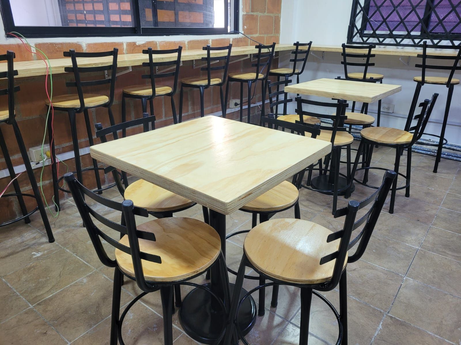 Cafetería vacía con mesas y taburetes de madera cerca de una ventana, dentro de una habitación con azulejos.