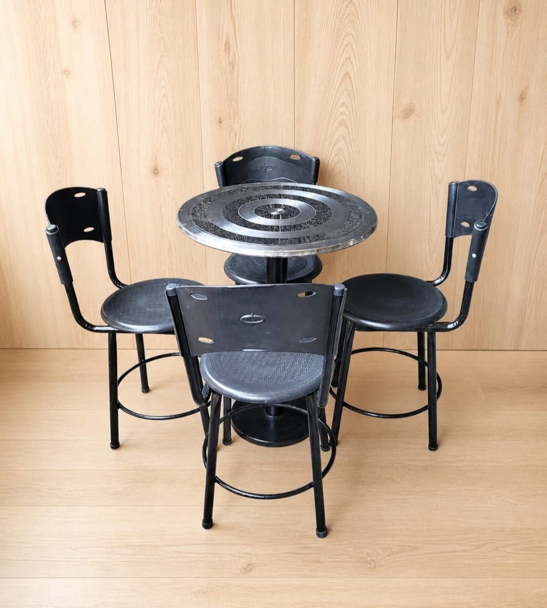 Mesa de patio de metal negro con cuatro sillas sobre un suelo de madera clara, contra una pared de madera.