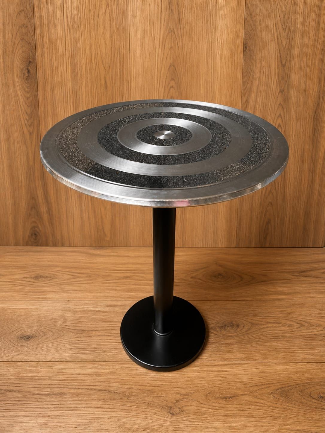 Mesa redonda de pedestal de metal con base negra, apoyada sobre una pared y un suelo de madera.