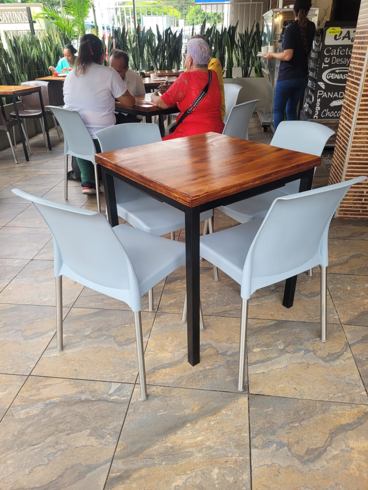 Mesa de café al aire libre con cuatro sillas claras y dos personas sentadas al fondo.