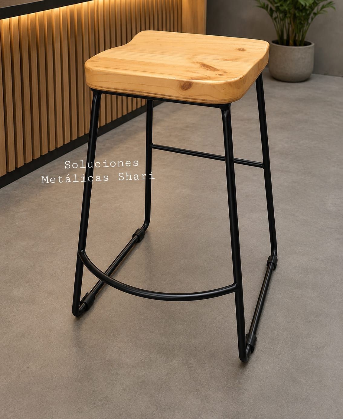 Taburete de bar con asiento de madera y patas de metal negro sobre un suelo gris, junto a una pared revestida de madera.
