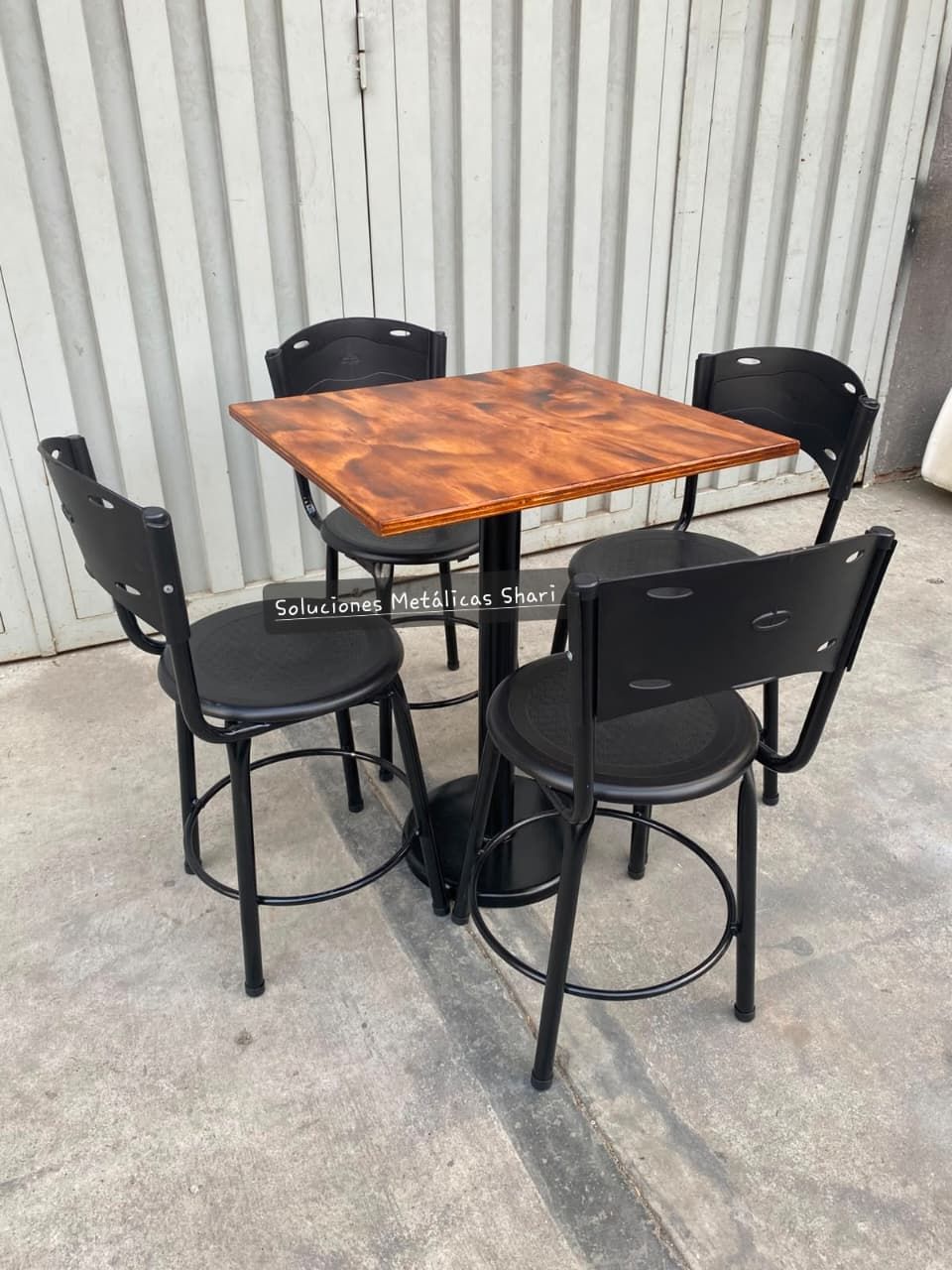 Mesa cuadrada de madera con cuatro sillas negras sobre un suelo de hormigón contra una pared de chapa ondulada.