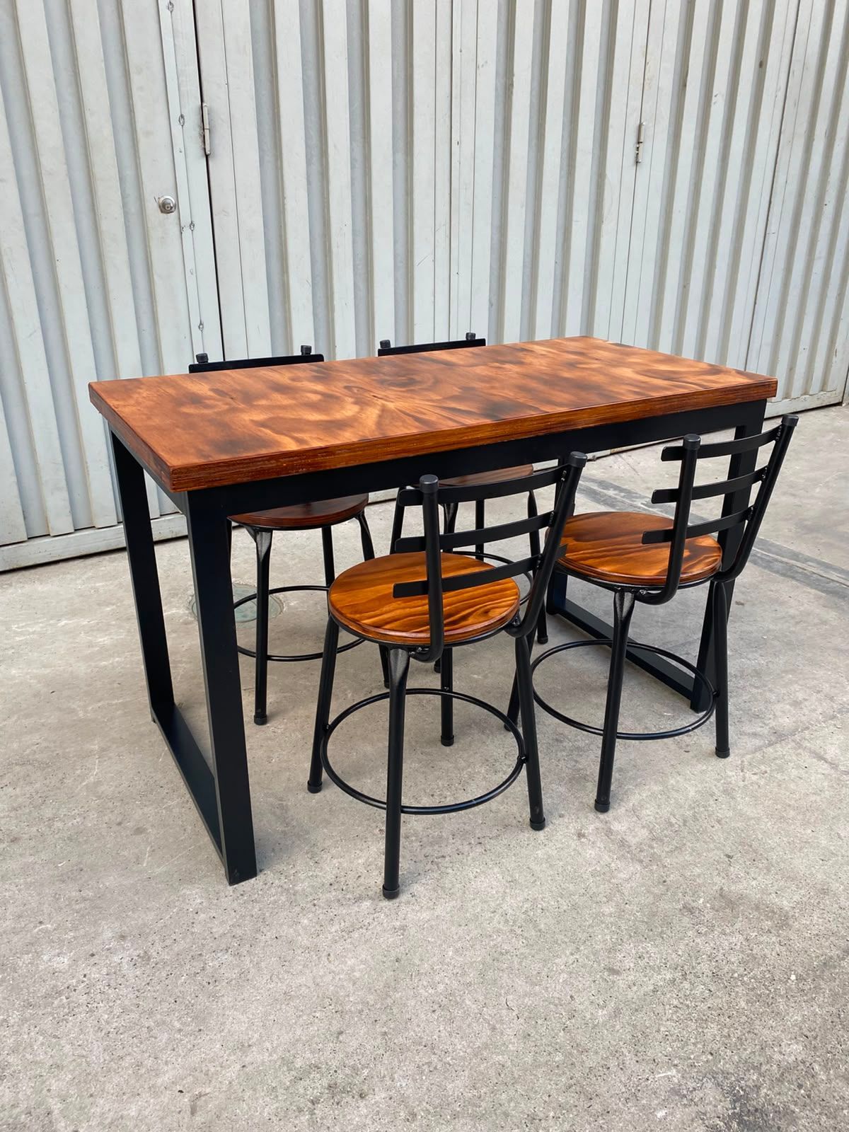 Mesa alta con tablero de madera y dos taburetes negros sobre un suelo de hormigón contra una pared de chapa ondulada.
