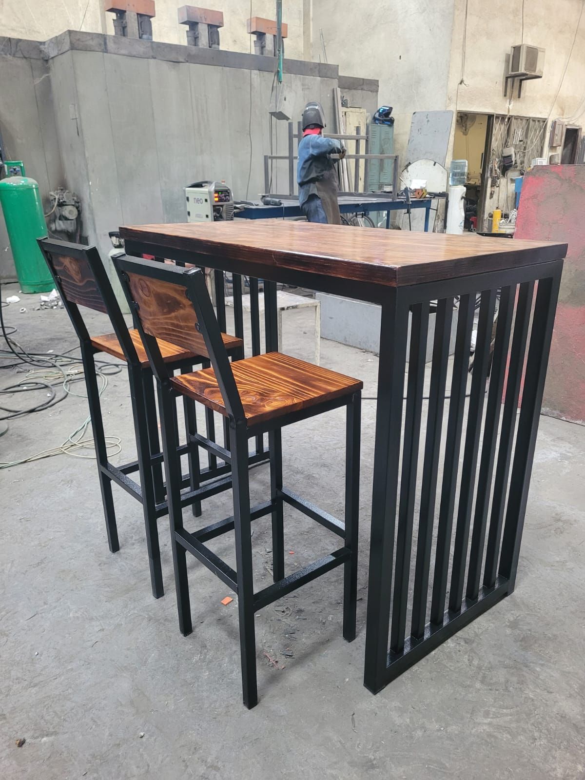 Mesa alta de madera con dos taburetes en una zona industrial al aire libre.