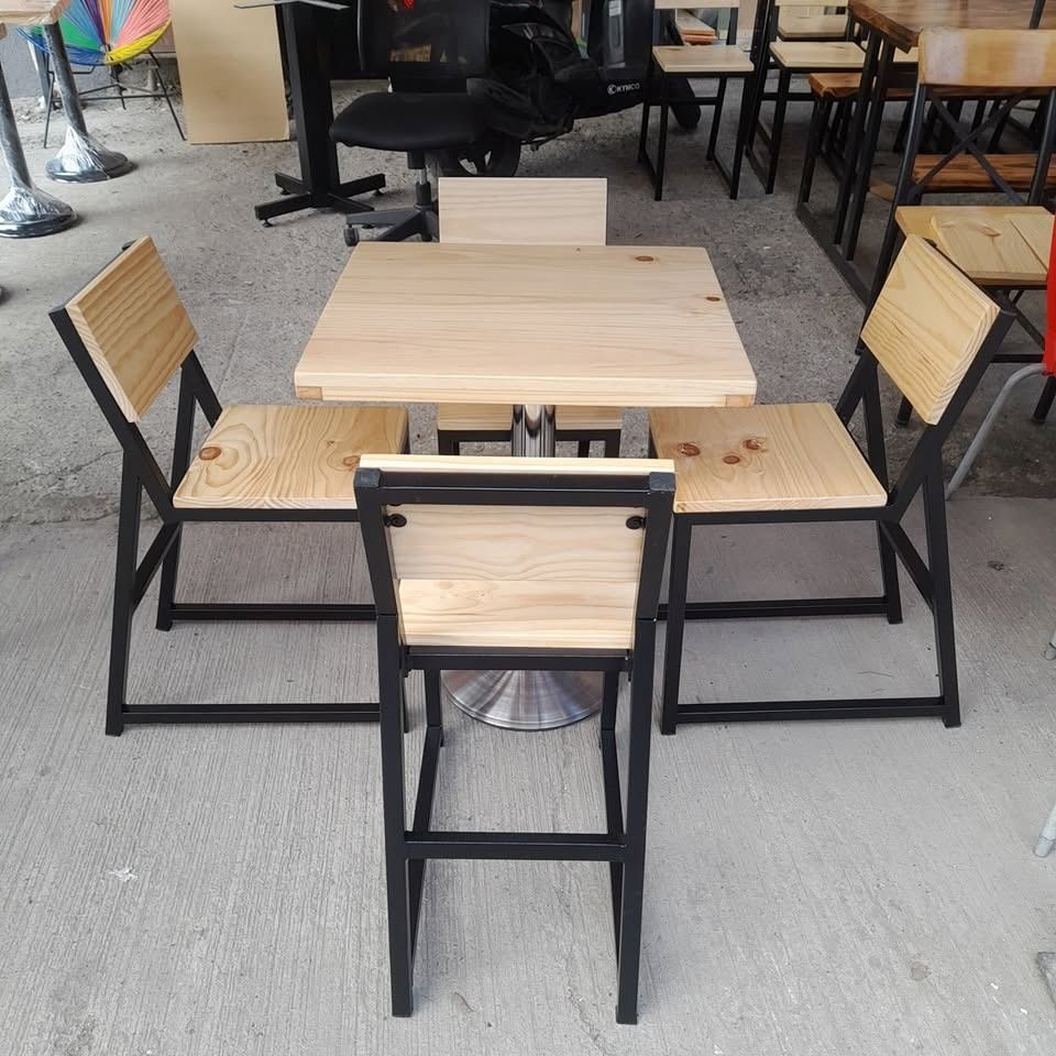 Mesa de café al aire libre con cuatro sillas de metal negro y tableros de madera clara sobre un suelo de hormigón.