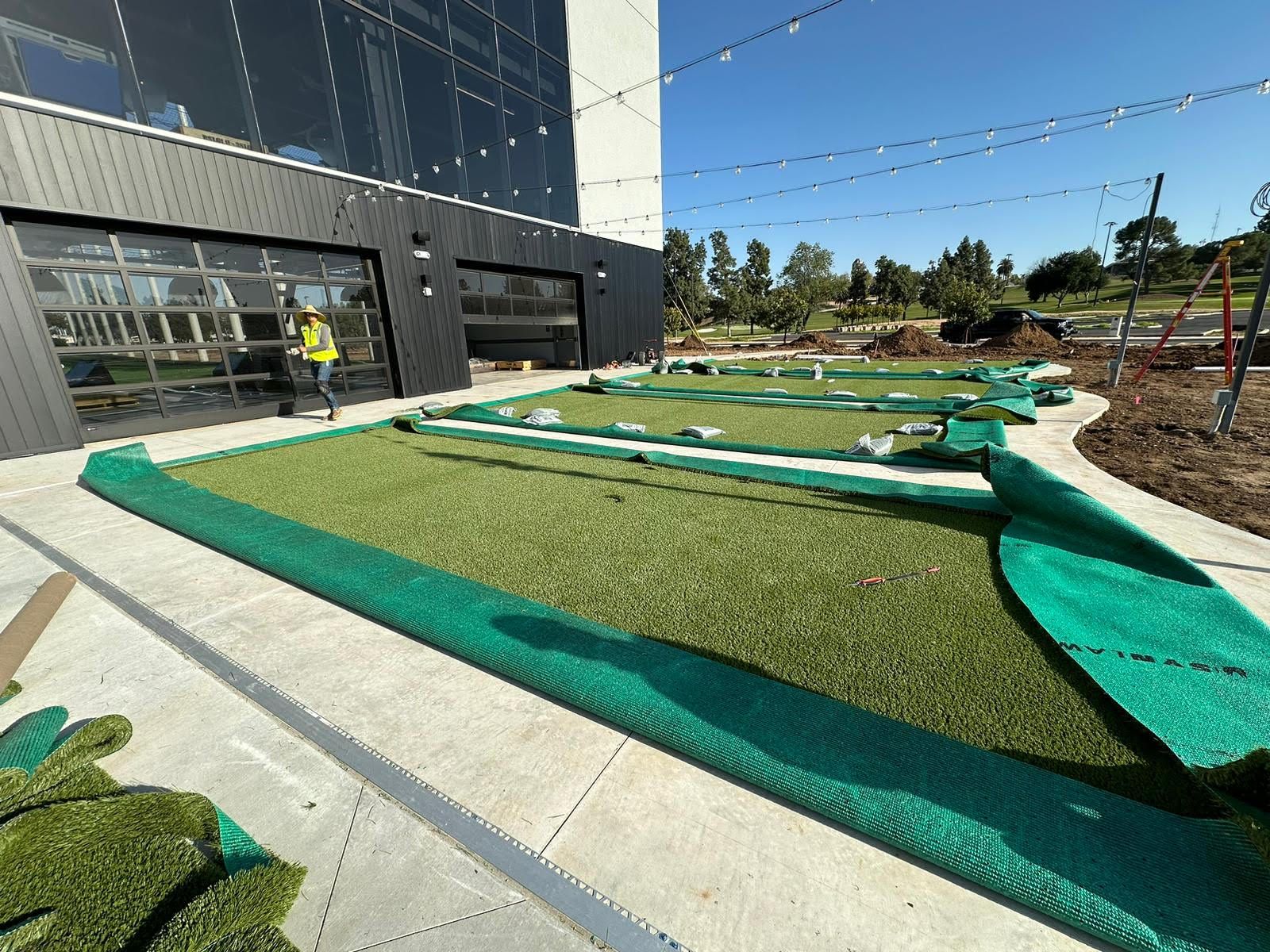 A worker walks near a modern building where green synthetic turf is being installed in outdoor strips.