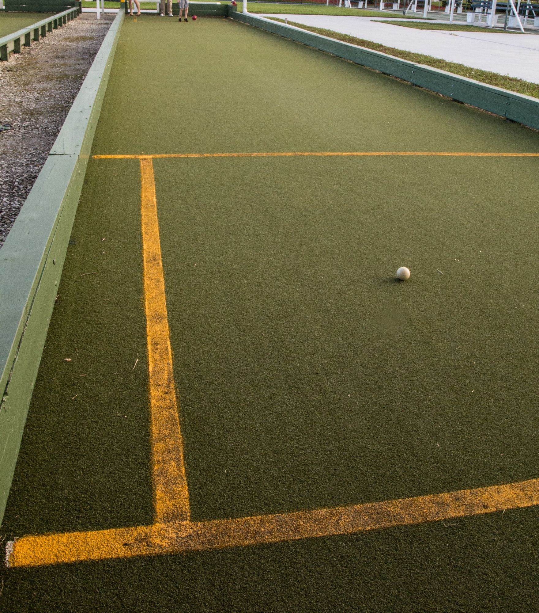 A white ball sits on a green bowling green next to yellow boundary lines, with wooden edges visible.