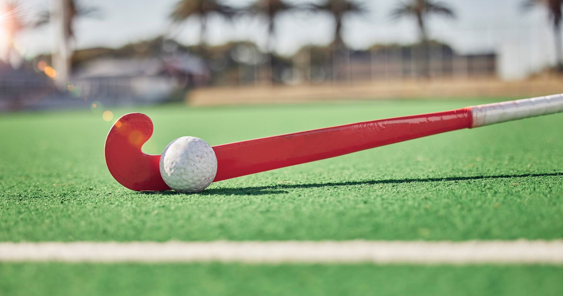 A red field hockey stick positioned against a white ball on a green synthetic turf field under a bright, sunny sky.
