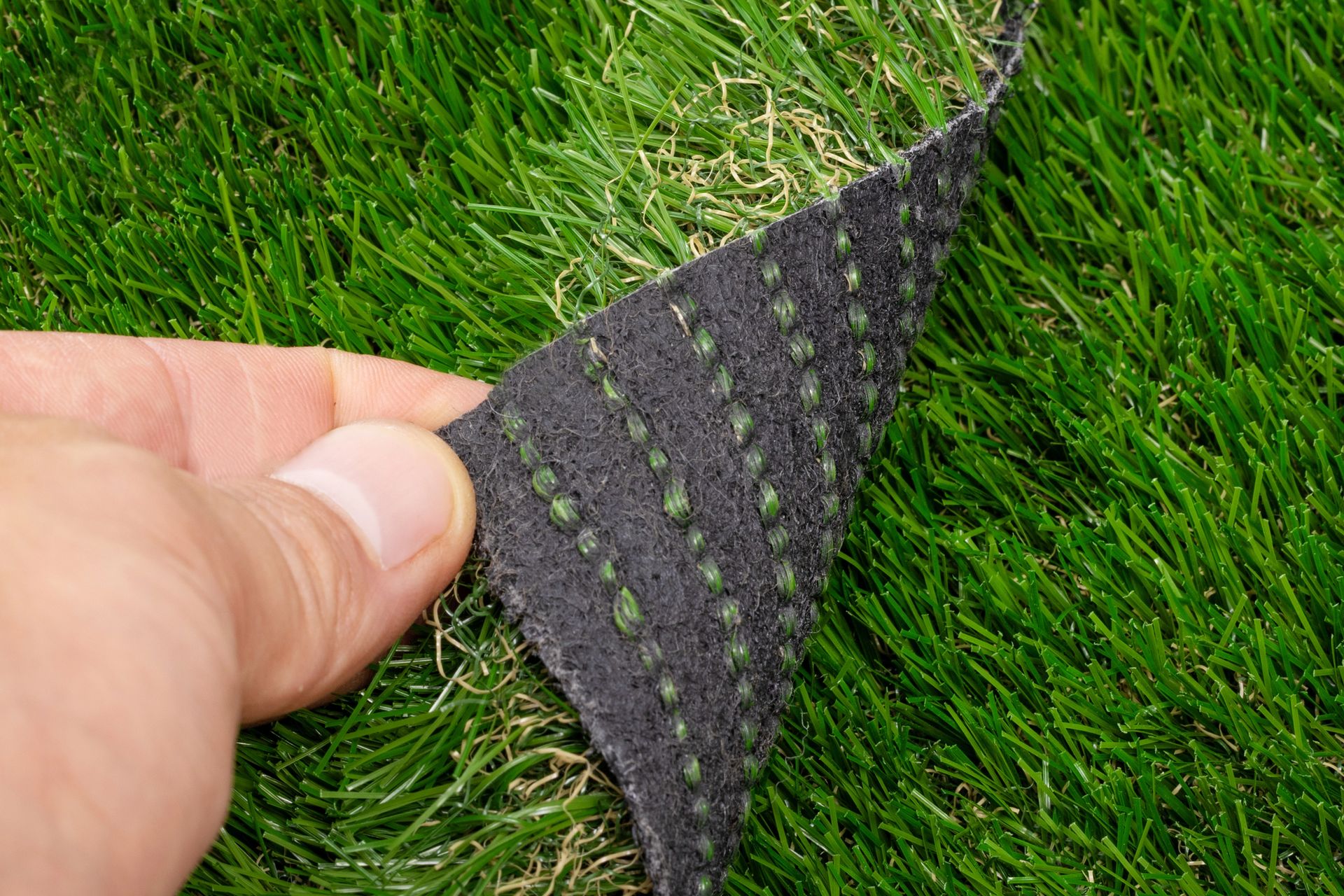 A hand lifts a corner of synthetic grass, revealing the dark, perforated backing with stitched rows of green fiber.