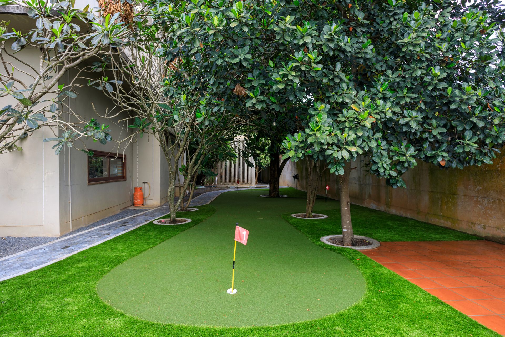 A backyard putting green with a red flag, set between a light-colored building and trees on a manicured lawn.
