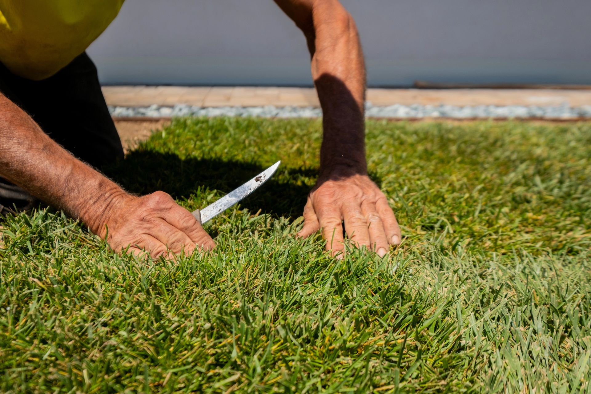 A person kneeling on a lawn uses a small knife to trim the edges of newly laid grass turf.