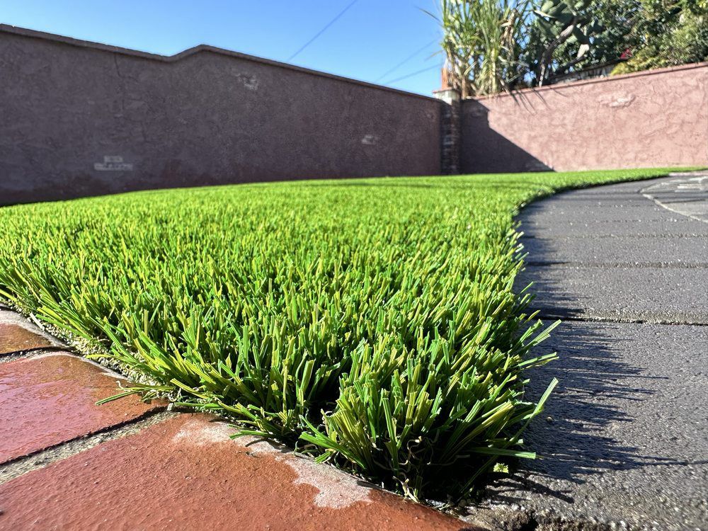 A close-up view of vibrant green artificial turf bordered by red bricks and dark grey pavers against a stucco wall.