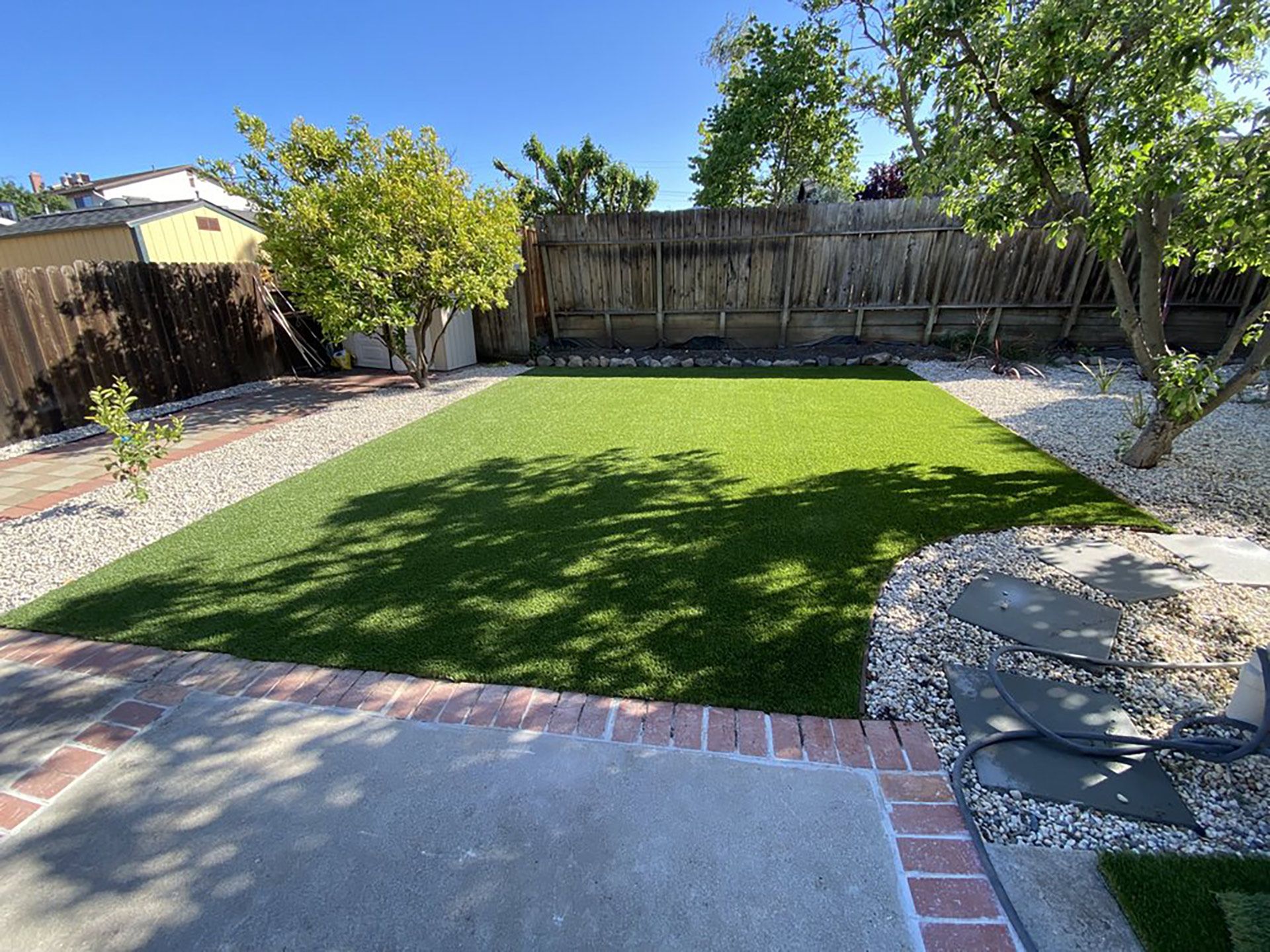 A rectangular patch of green artificial turf in a backyard, framed by light-colored gravel, trees, and a wooden fence.