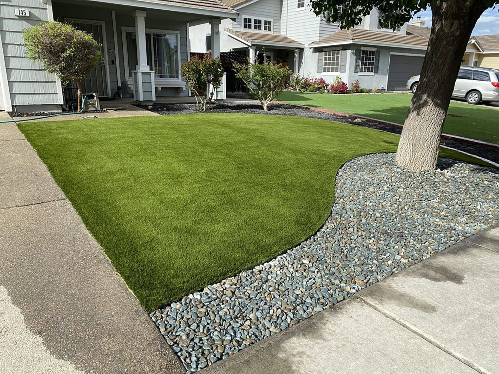 A neat front yard featuring a patch of green grass bordered by decorative gravel and a large tree next to a house.